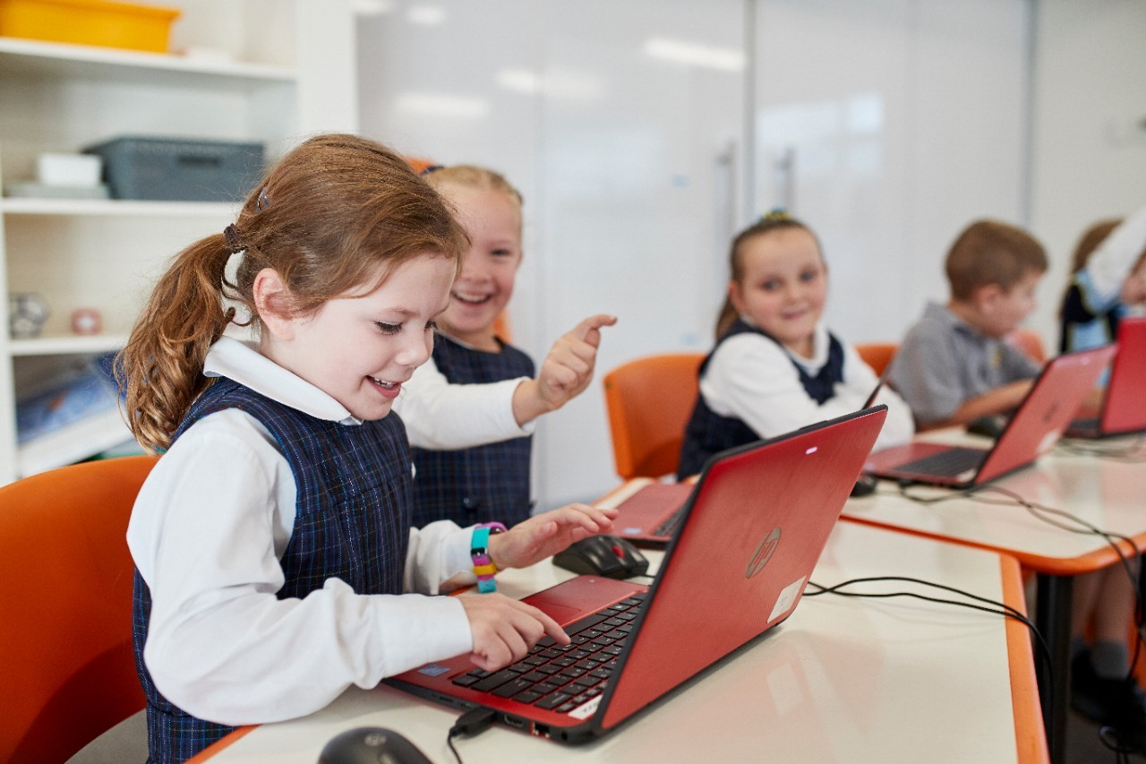 Primary_school_students_using_computers_in_classroom_2_-_Web_Optimized_Image Students sitting ad a desk and typing on a red laptop.